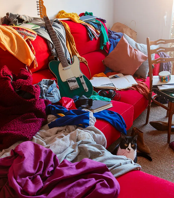 A cluttered red couch in someone's home piled high with clothes, books, and a guitar
