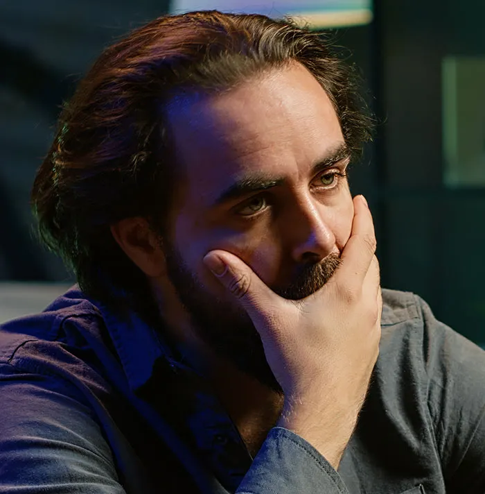 A concerned bearded man holding his chin while inside someone's home.