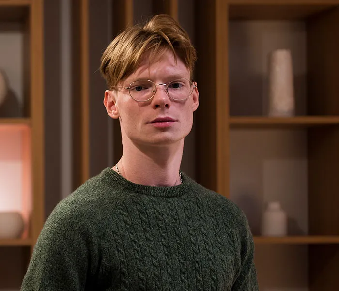 A young man with glasses and a green sweater standing in a home interior