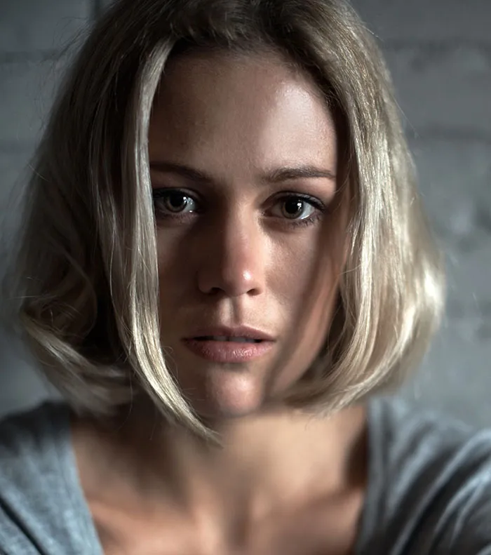 A serious woman with short blonde hair looking directly at the camera indoors