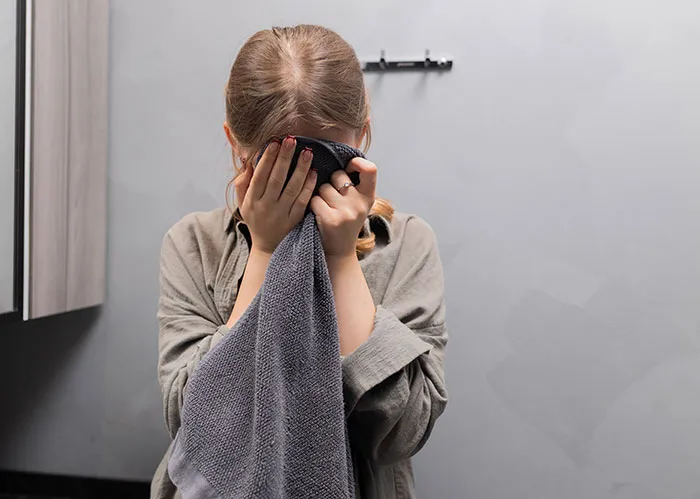 A woman drying or hiding her face with a gray hand towel in someone's home bathroom.