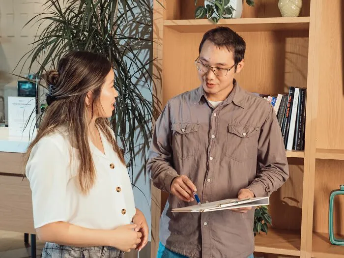A man with glasses holding a clipboard talking to a woman in a tiny community office.