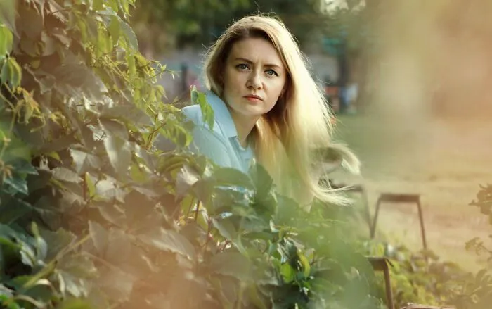 A blonde woman looking attentively through green leaves as if trusting her gut feeling.