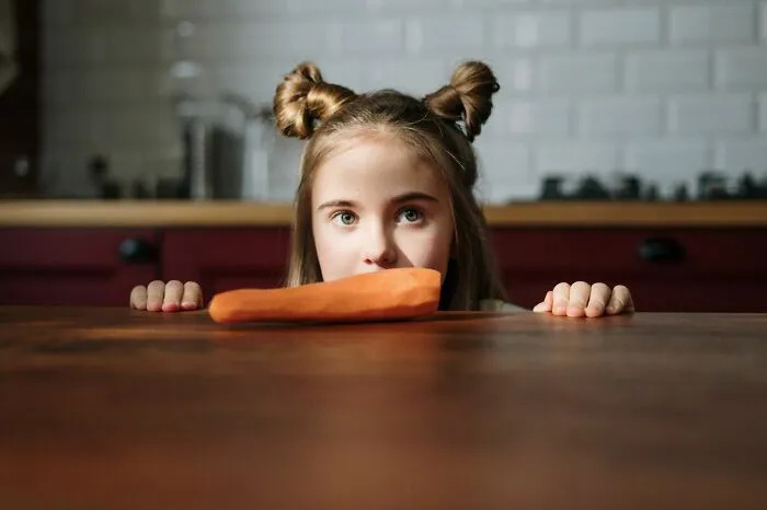 A young girl looking over a table at a raw carrot, expecting candy while trick-or-treating but receiving veggies.