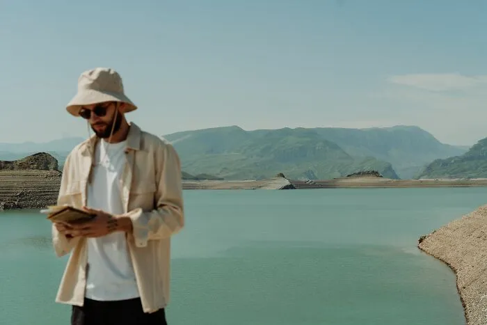 A man wearing a bucket hat and holding a notebook with a scenic lake and mountains behind him.