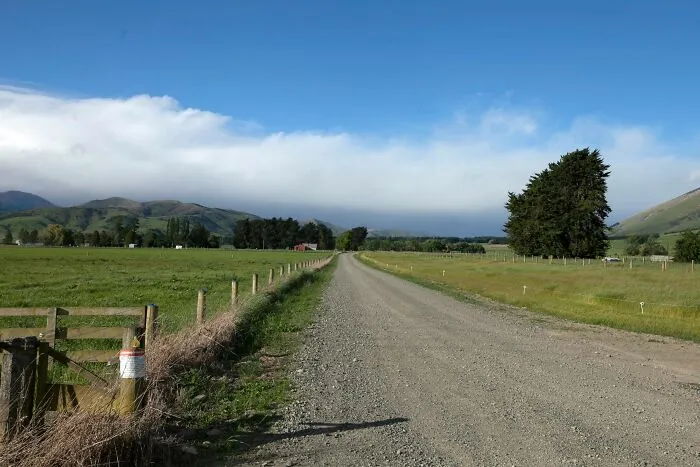 A long gravel road passing through green fields in tiny communities with mountains in the distance