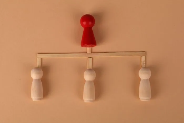 Red wooden game piece positioned above three plain wooden figures on a structure representing corporate structure in companies