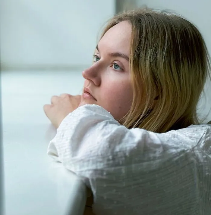 A blonde woman leaning her head on a surface and looking pensively out a window, burdened by secrets.