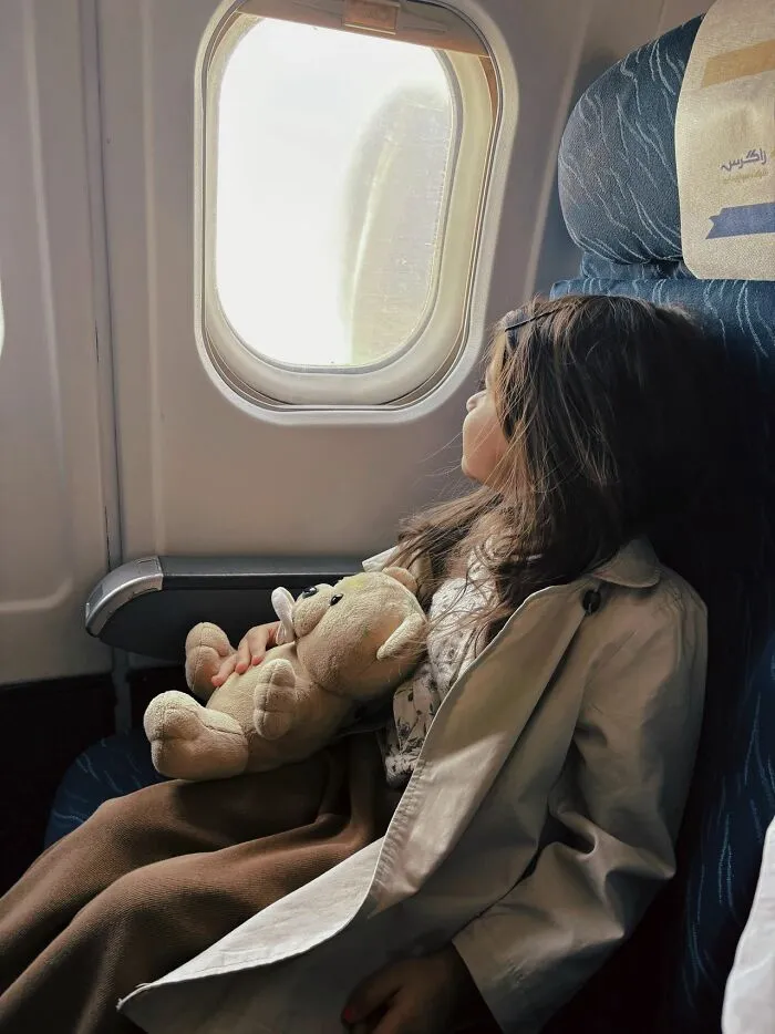 A young girl holding a teddy bear looking out the window to get better seats on flights