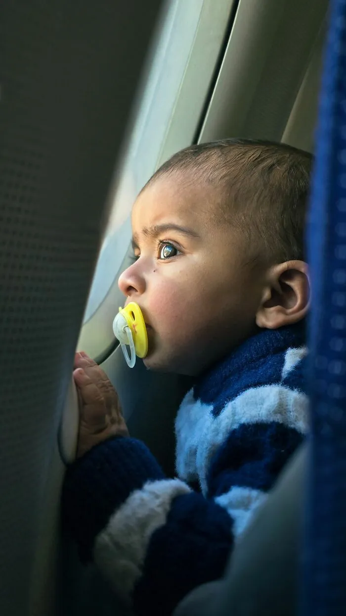 A baby with a pacifier looking out the window to get better seats on flights