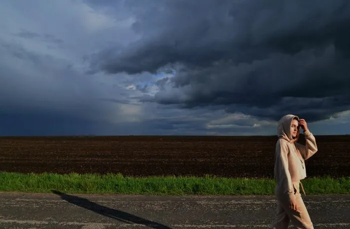 A girl in a hoodie walking by a field under dark storm clouds, sensing an urgent feeling to leave