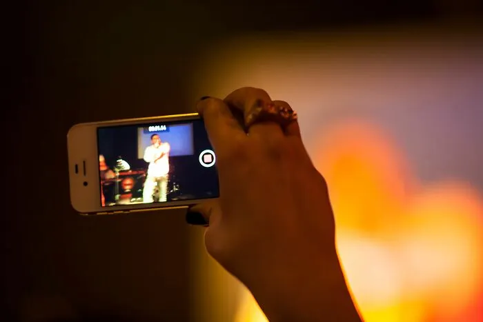 A hand holding a smartphone to record a live performance on screen, depicting unethical life pro tips