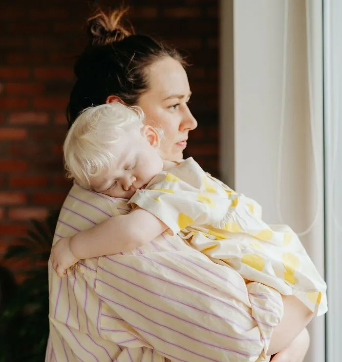 A mother holding her sleeping child near a window, pondering life secrets.