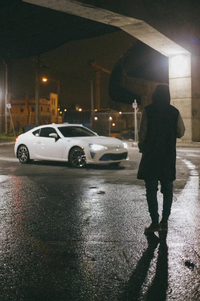 A person standing by a white car at night, evoking the urgent feeling of needing to leave immediately