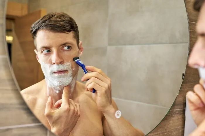 A young man shaving his face with foam in a bathroom mirror, demonstrating everyday clever life tricks for grooming.