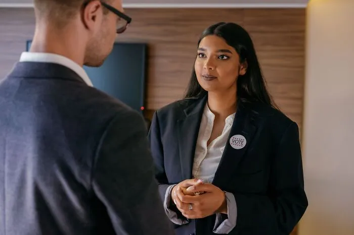 A woman in a blazer speaking to a man wearing glasses.