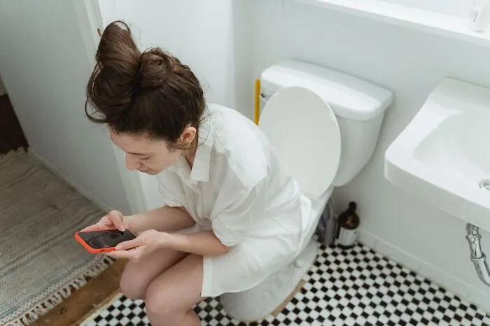 A woman sits on a toilet using a smartphone amidst misconceptions about women's bodies.