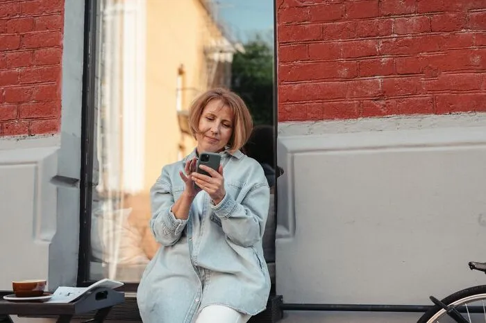 Middle aged woman sitting outdoors and looking at her smartphone during funny instances.