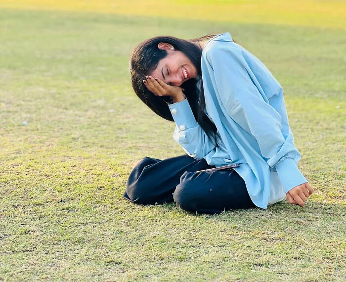 A young woman laughing embarrassingly while sitting on the grass outdoor.