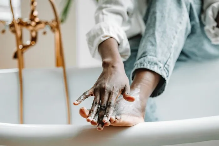A person applying moisturizer to their foot while sitting on the edge of a bathtub.