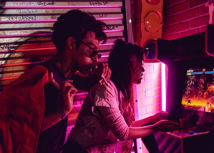 Two young people playing an arcade game under neon lights, evoking chilling stories.