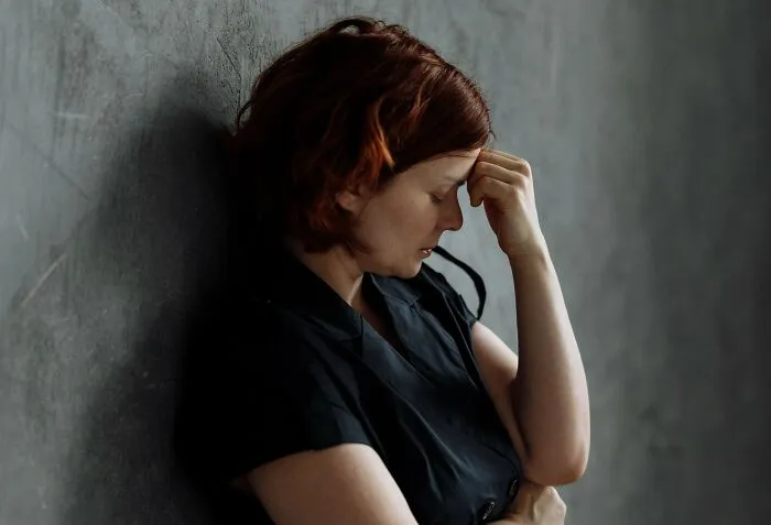 A red-haired woman leaning against a wall with her hand on her forehead, stressed about secret burdens.