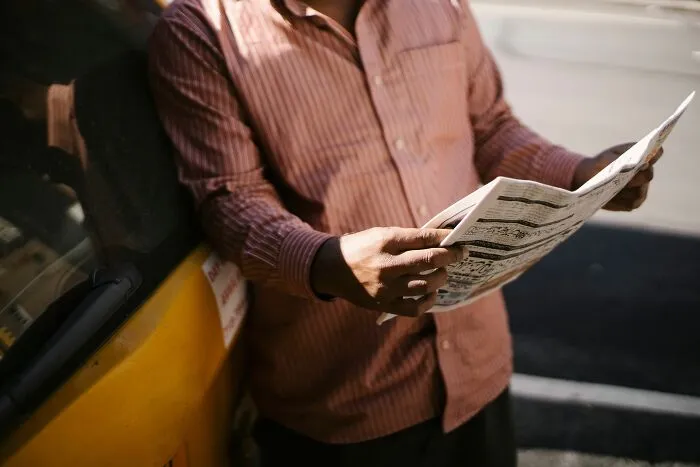 Man reading a newspaper beside a yellow car, catching up on tiny communities drama
