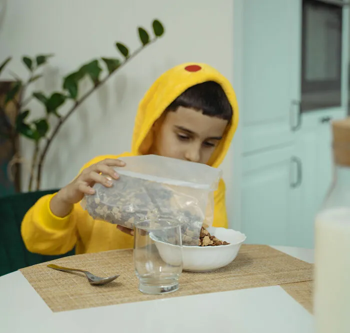 Child in yellow hoodie pouring cereal into bowl at kitchen table