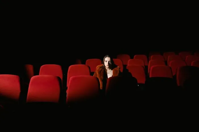 Woman sitting alone in an empty dark movie theater showing red seats, illustrating funny instances.
