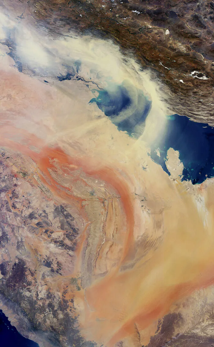 A surreal satellite photo capturing a vast dust storm blowing across desert terrain and over a body of blue water.