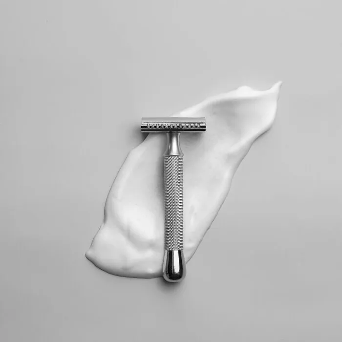 A classic metal safety razor placed over white shaving cream on a gray background