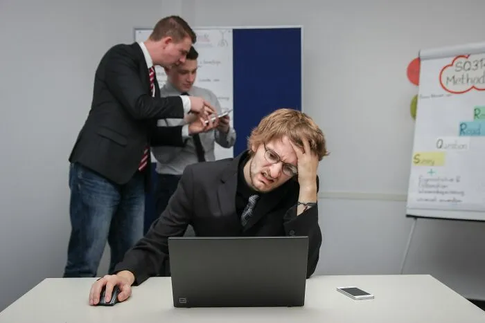 A stressed man working at a laptop while colleagues talk, illustrating how professionals lose trust.