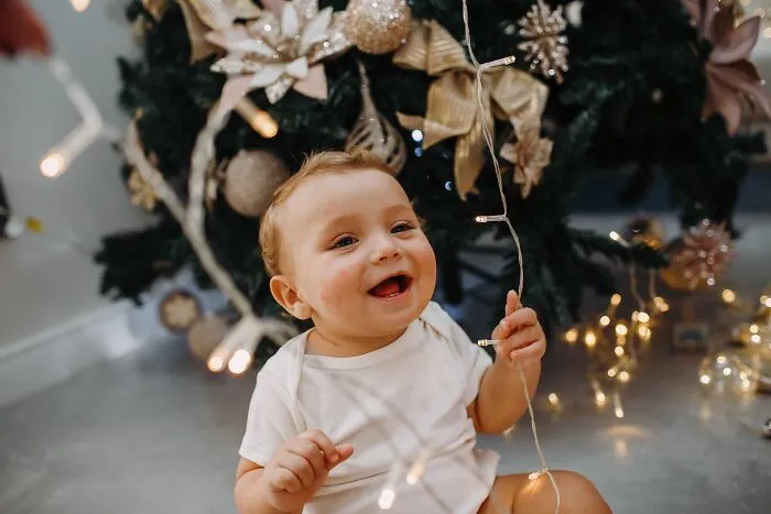 Smiling baby holding Christmas lights near decorated tree