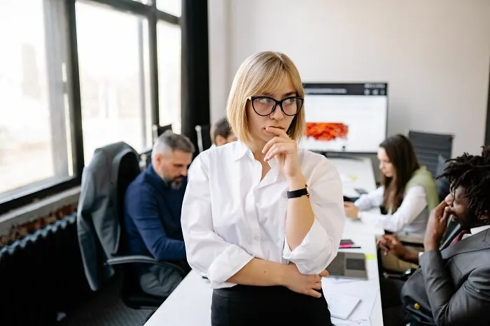 Thoughtful woman in glasses standing in office with coworkers
