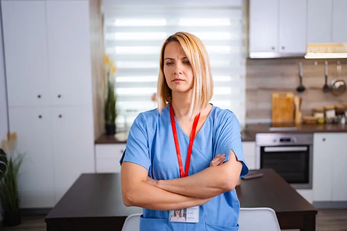 A blonde female healthcare worker in blue scrubs with arms crossed looking serious.
