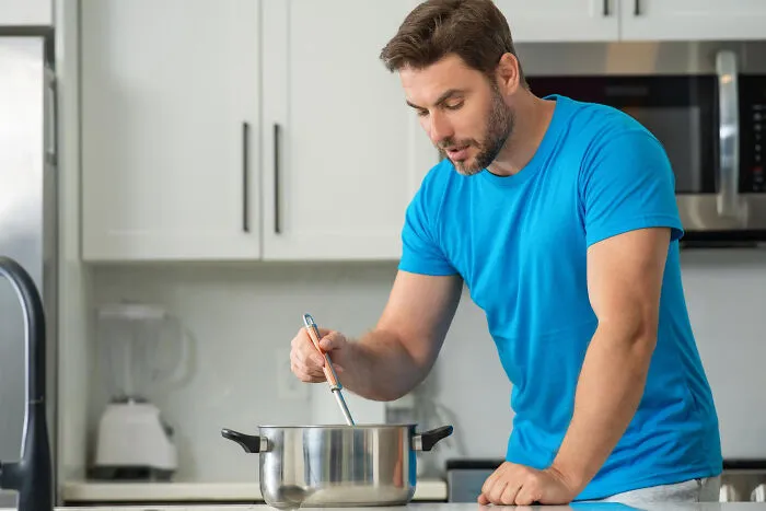 A young man in a blue t-shirt cooks and stirs a pot in a bright kitchen, showcasing life living with guys.
