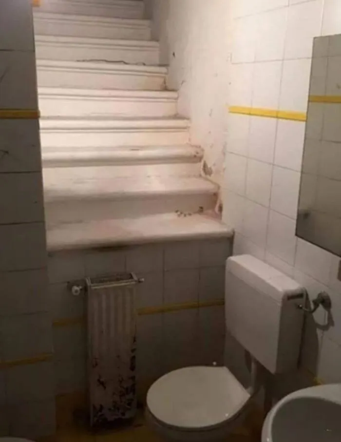 Escalera instalada dentro de un baño pequeño con inodoro y radiator viejo, ejemplo de personas que menos mal que hicieron una foto.
