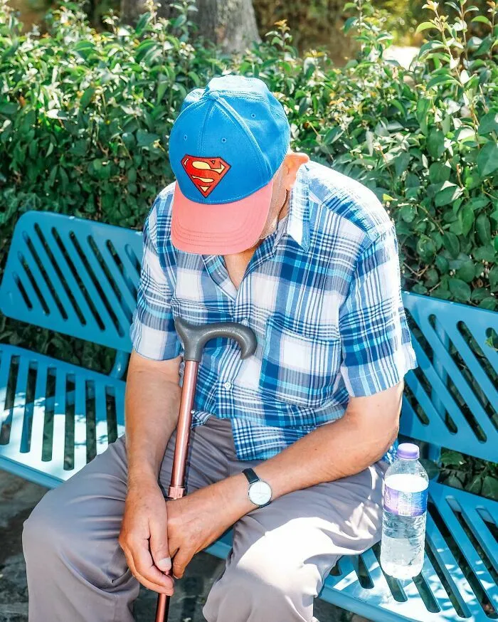 Anciano descansando en un banco del parque con gorra de Superman en curiosas escenas callejeras.