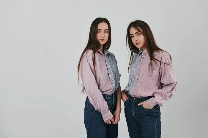 Dos jóvenes gemelas idénticas posando juntas con camisas similares sobre fondo gris.
