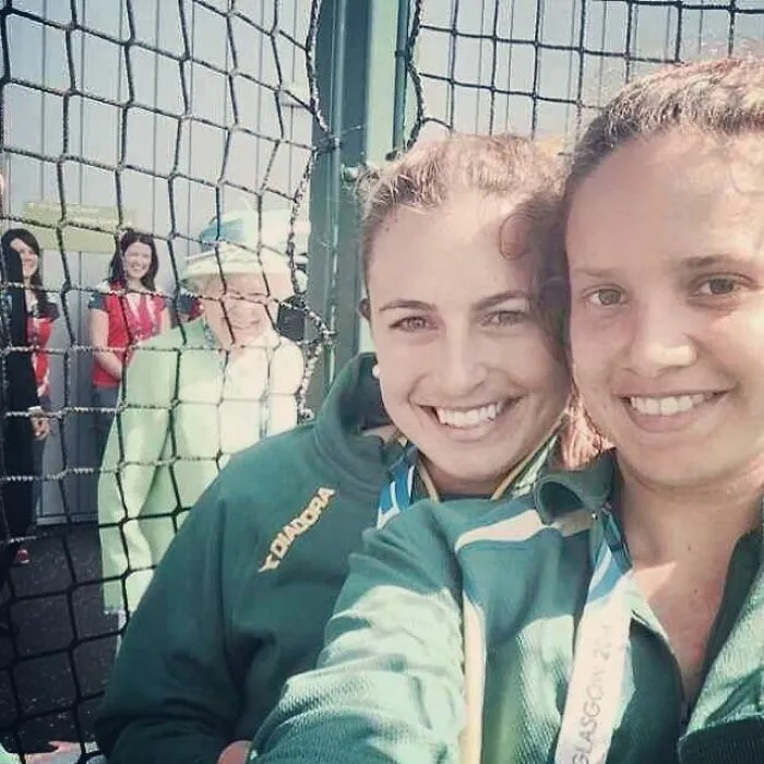 Divertidas fotos con la Reina Isabel sonriendo en el fondo detrás de dos jóvenes en primer plano.