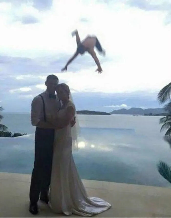 Novios posando junto al mar mientras alguien salta de cabeza al fondo de divertidas fotos de boda.