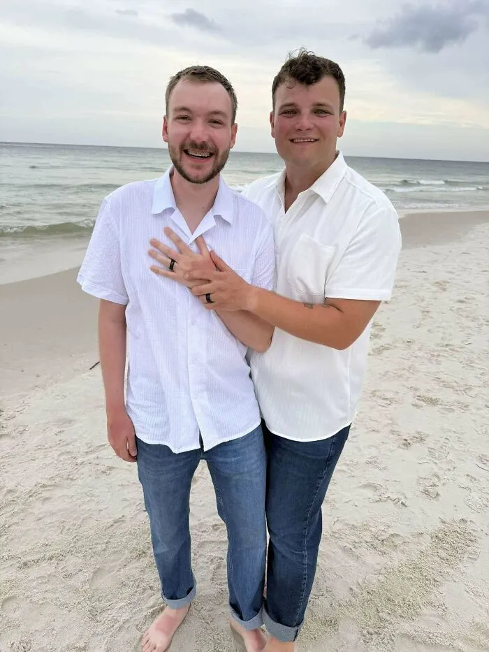 Pareja sonriente abrazada en la playa mostrando sus anillos, una de las publicaciones reconfortantes del día.